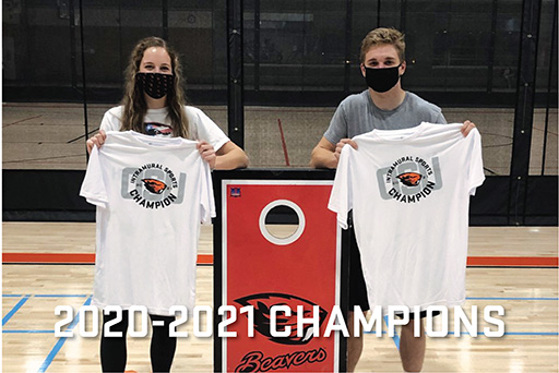 Photo with text overlay that says 2020-2021 Champions. Photo shows two masked participants with cornhole board between them showing their champions t-shirts.