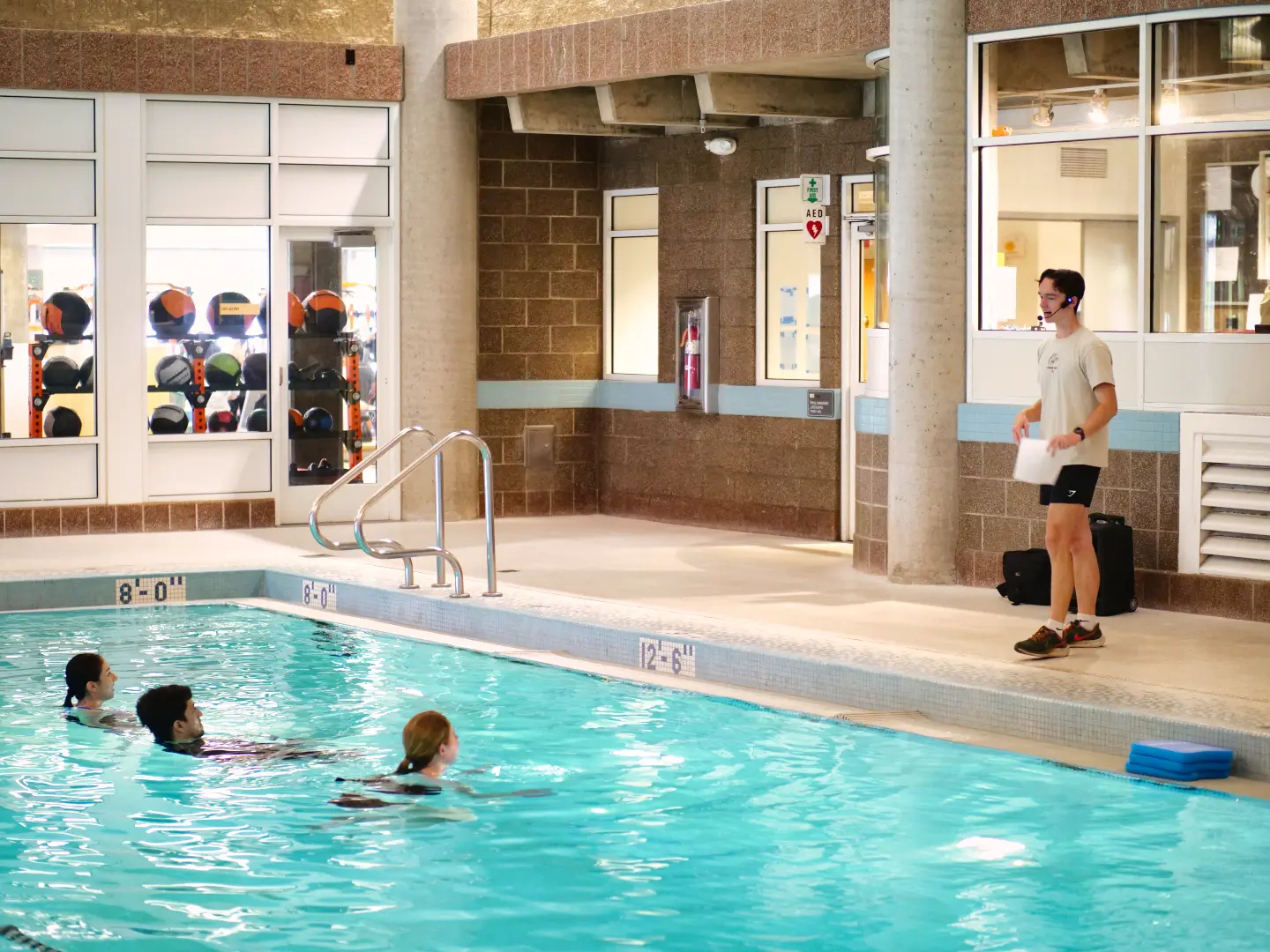 An indoor swimming pool with several people exercising in the water as an instructor stands at the pool’s edge holding a clipboard.